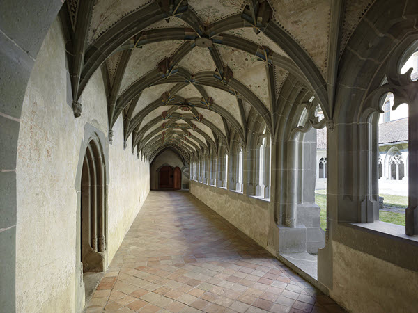 View of the southern part of the covered walkway. Viewing direction from east to west. On the left there is a wall with a door. The windows of the covered walkway and a door open onto the inner courtyard on the right. The floor is covered with fired tiles. A gothic rib vault covers the ceiling. The ribs of the vault and the borders of the windows and the doors are made of grey sandstone. Decorated plaster covers the triangle shapes above the arch. It’s daytime and the light is subdued.